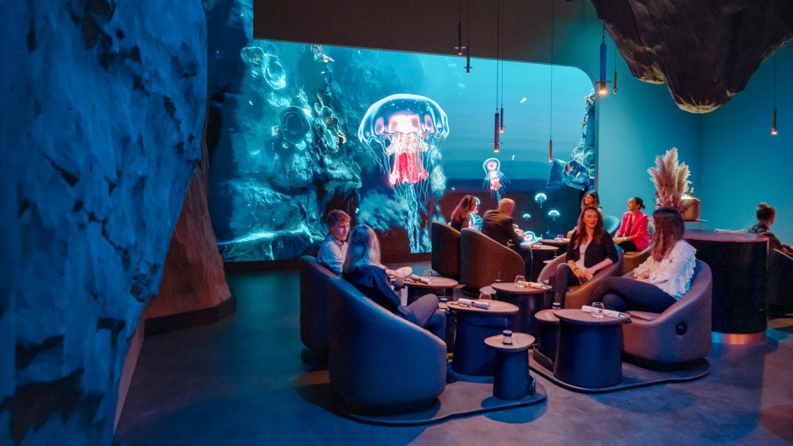 Gäste des Eatrenalin sitzen in modernen Loungesessel vor Couchtischen. Wände und Decke ähneln einer Unterwasser-Grotte. Eine Wand wird großflächig mit Unterwasserszenen samt leuchtender Quallen bespielt.