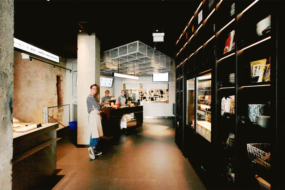 Ansicht des Mochi-Shops o.m.k: ein elegantes Holzregal mit Delikatessen und Zutaten der japanischen Küche. gegenüber Lehnt ein Mitarbeiter mit Schürze.