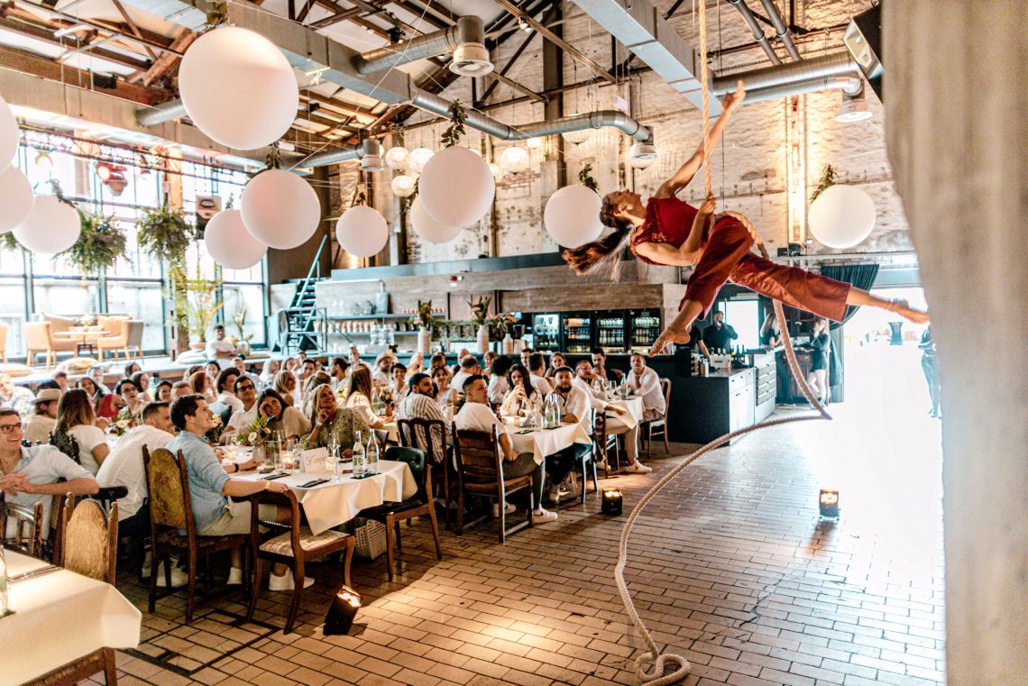 In der ehemaligen Siefenfabrikshalle reihen sich lange festlich gedeckte Tafeln unter Rohren und Stahlträgern. Eine Gruppe diniert und bestaunt eine Akrobatin, die kunstvoll in der Luft an einem Seil turnt.