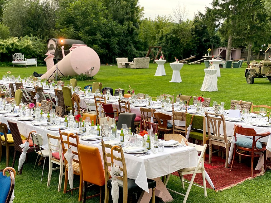 Garten der Stadtflucht Bergmühle: Auf einem Rasen stehen zwei lange festlich gedeckte Tafeln, die Sessel sind einen kunterbunte Mischung. Im Hintergrund taucht eine Metallskulptur eines rosa U-Boots scheinbar aus dem Rasen auf.