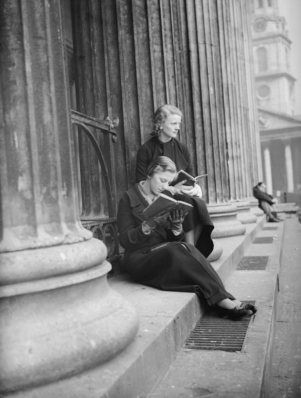 Zwei Frauen sitzen lesend auf den Stufen zwischen den Säulen eines sehr großen Gebäudes.