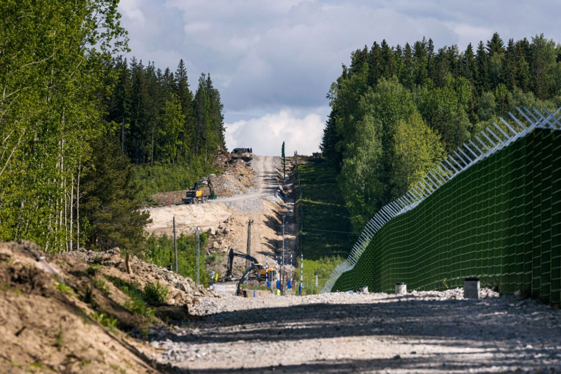 Ein Wald mit einem breiten gerodeten Streifen mit einem grünen Metallzaun und Baumaschinen. Der Zaun wird von Finnland an der Grenze zu Russland errichtet.
