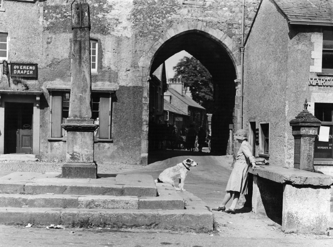 Ein Hund sitzt auf der Treppe eines Denkmals und blickt auf eine Frau mit Hut, die ihm gegenüber an einem Geländer lehnt. Die schwarz-weiß Aufnahme entstand in den 1930 Jahren. Das Bild illustriert einen Beitrag über Spitzengastronomie und Haubenküche.