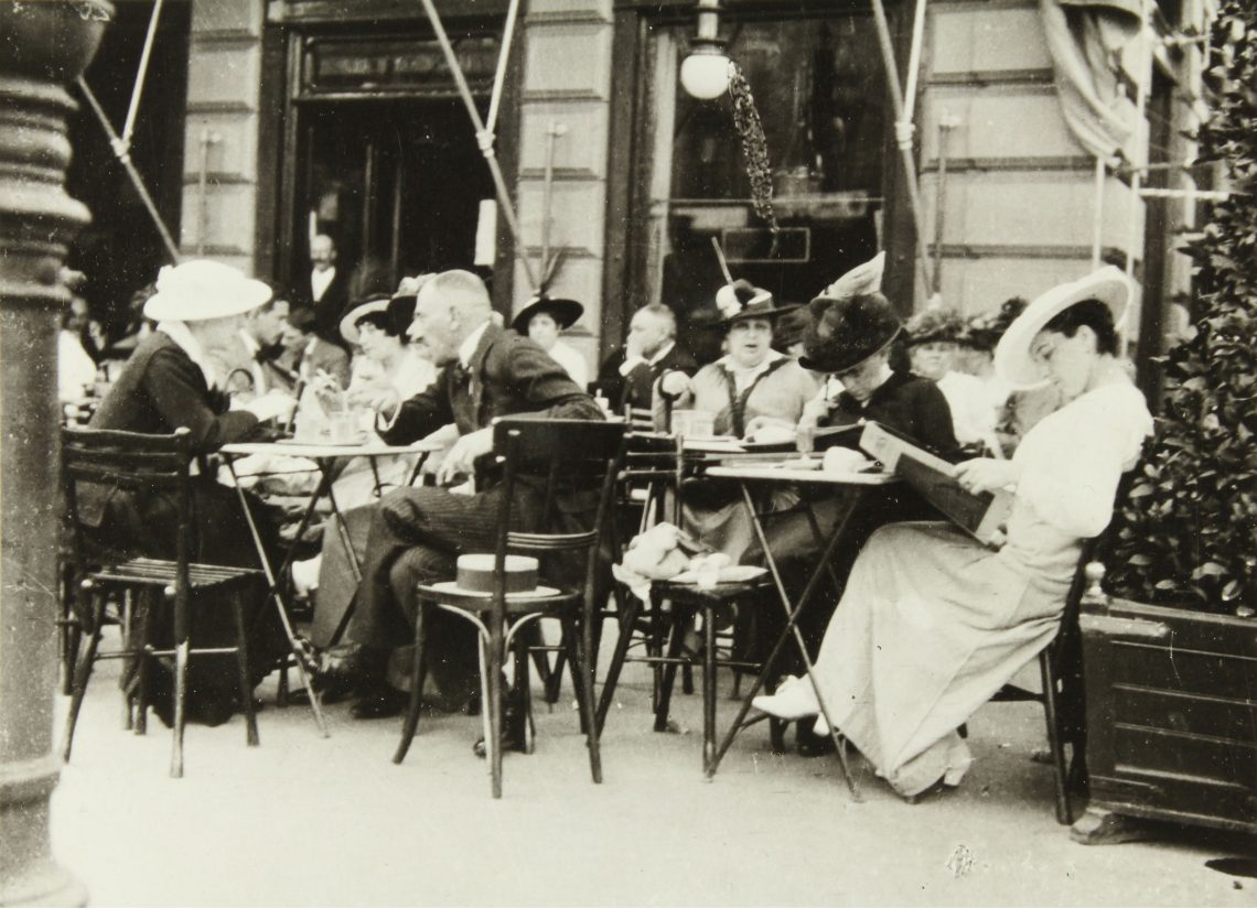 Gäste eines Restaurants oder Kaffeehauses sitzen sich unterhaltend oder lesend an Tischen in einem Schanigarten.