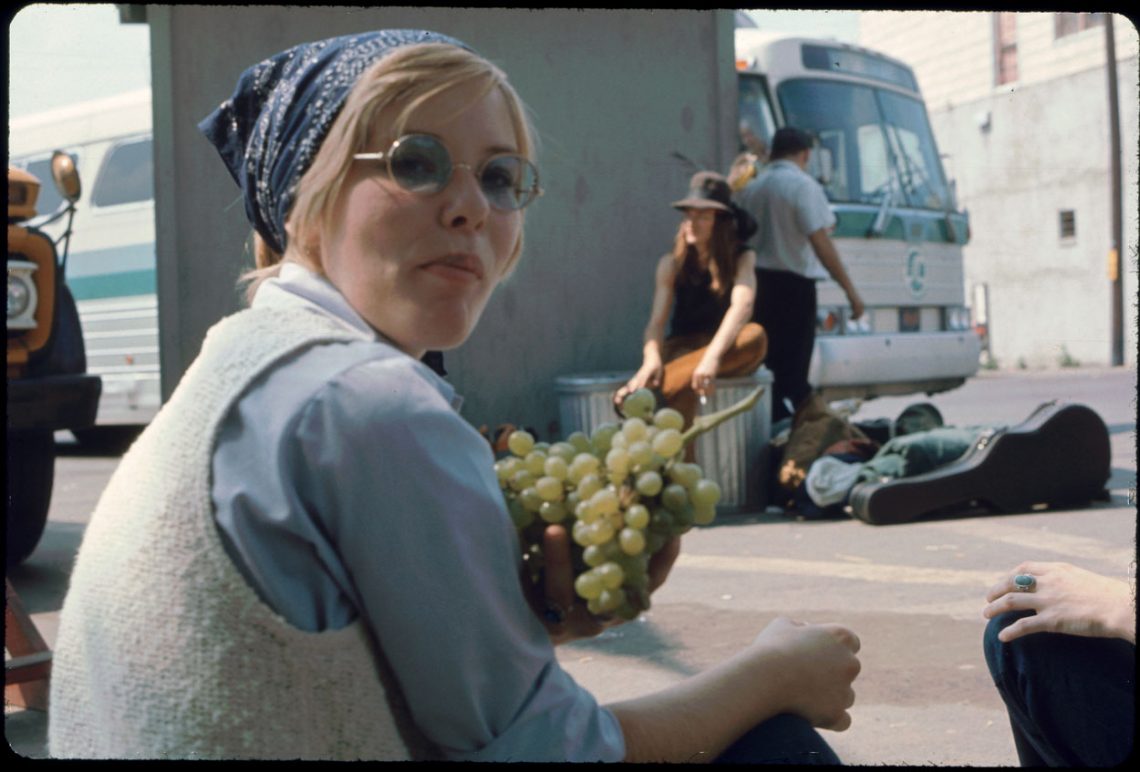 Foto einer jungen Frau auf einem Busbahnhof, die Trauben isst und über die Schulter in die Kamera blickt.