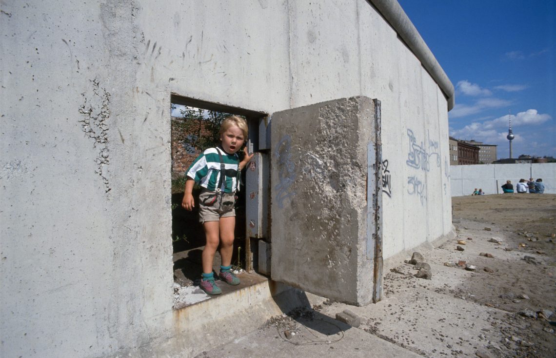 Ein kleiner Junge steht in einer Öffnung der Berliner Mauer, die wie eine Tür aus dieser herausgeschnitten ist. Er trägt eine Lederhose und ein grün-weiß gestriftes T--Shirt. Das Bild ist Teil eines Beitrags über Europa und die Globalisierung, die mit dem Fall der Berliner Mauer und später durch den Zerfall der Sowjetunion noch stärker wurde.