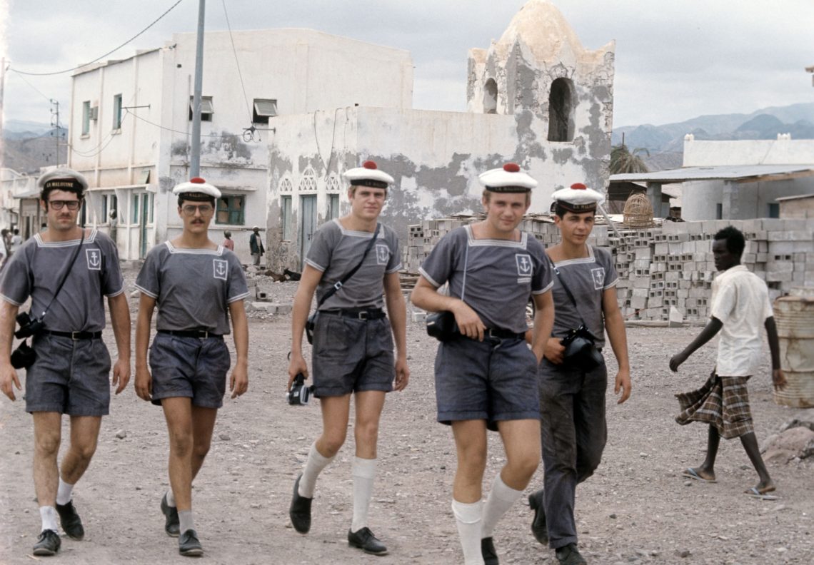 Eine Gruppe Männer mit weißen Mützen mit rotem Bommel geht eine Straße entlang. Im Hintergrund betrachtet ein Mann die Gruppe scheinbar irritiert.