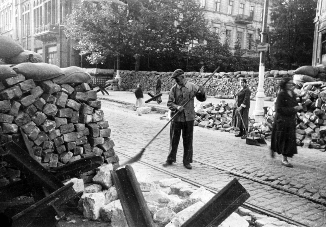 Ein Mann und eine Frau fegen eine Straße, die mit Sandsäcken gesäumt ist, während eine weitere Frau vorbeigeht. Im Hintergrund sind Gebäude aus der Zeit um 1900 zu sehen. Das Bild ist Teil eines Beitrags über die Ukraine und die Getreideexporte. Das Foto wurde in Odessa aufgenommen, 1941.