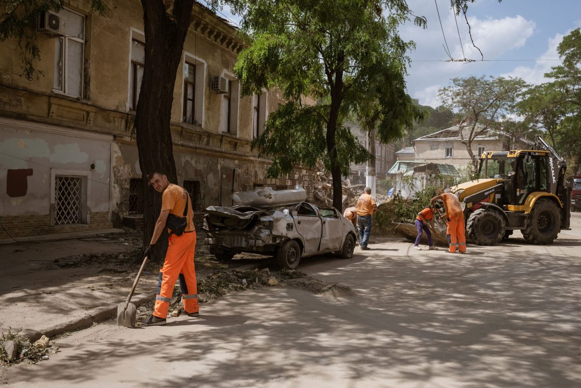 Ein Mann in organger Kluft reinigt mit einer Schaufel eine Straße von Schutt. Im Hintergrund steht ein zerstörtes Auto und ein Bagger mit dem weiterer Schutt abtransportiert wird. Die Aufnahme ist in Odessa.