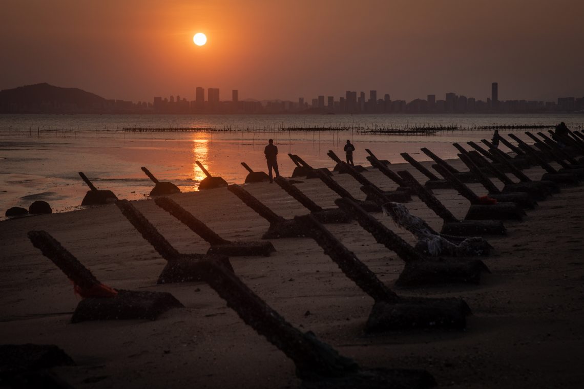 Ein Mann fotografiert den Sonnenuntergang über der chinesischen Stadt Xiamen von der taiwanesischen Insel Kinmen aus. Panzersperren von vergangenen Konflikten säumen den Strand. Das Bild illustriert einen Beitrag zu Europa im globalen Machtkampf.