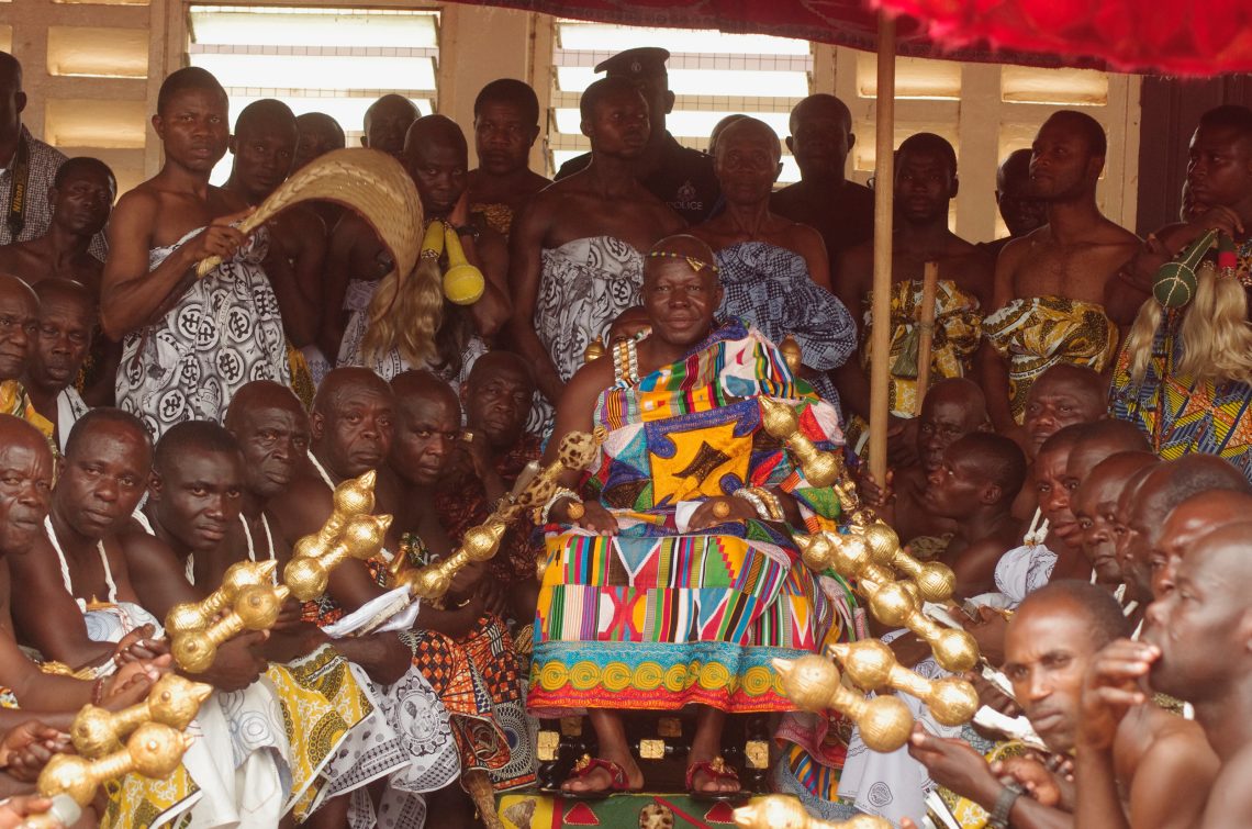 Der König der Ashanti in Ghana Osei Tutu II sitzt bunt gewandet umgeben von Mitgliedern seines Hofs, die goldene Szepter halten. 