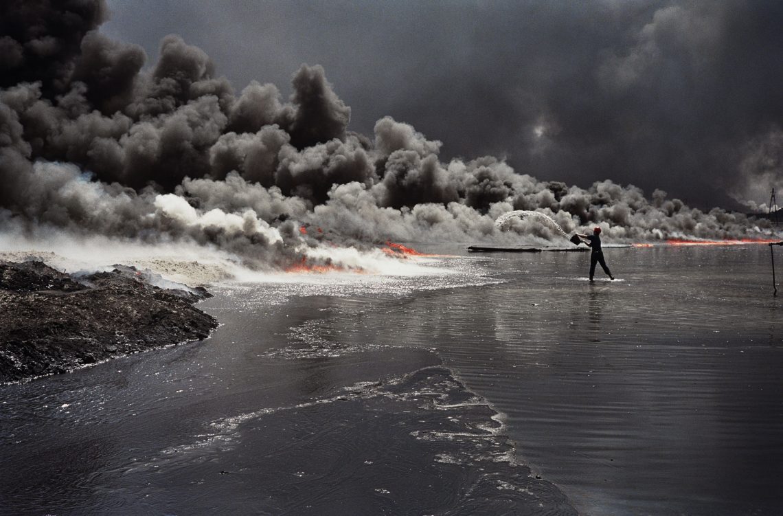 EIn Mann steht im Wasser einer riesigen Rauchwolke gegenüber, mit einem Eimer schüttet er Wasser in die Flammen. Das Bild zeigt brennende Ölfelder in Kuweit.
