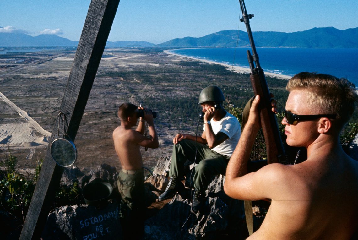 Drei Soldaten, einer mit Fernglas, einer mit einem Gewehr, einer telefonierend sitzen auf einem Aussichtspunkt. Unter ihnen eine Ebene mit Meer und Strand. Das Bild wurde 1965 in Vietnam aufgenommen und ist Teil eines Beitrags über die Klimafolgend des Kriegs in der Ukraine.