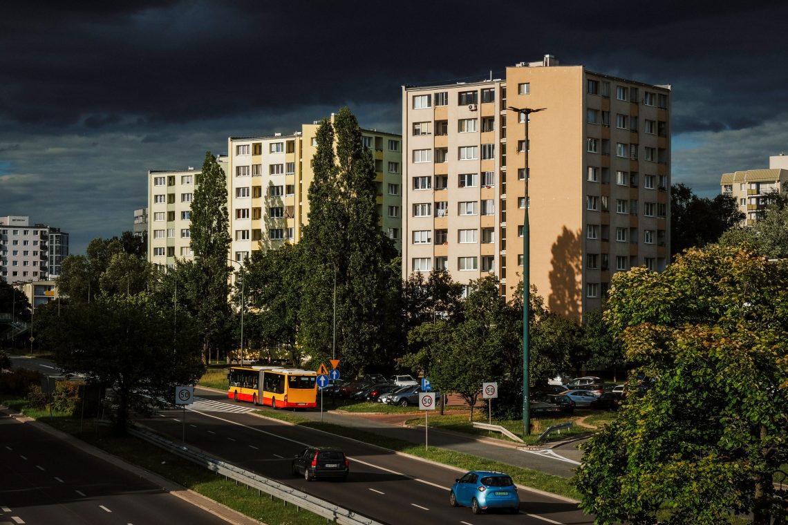 Abendlicht auf einer Straße mit Wohnblöcken aus der Sowjetzeit in Warschau, Polen. Vor den vor einem dunklen Himmel leuchtenden Wohnblocks ist eine breite Straße mit Bäumen und ein Bus. Das Bild illustriert einen Beitrag über Zinsbremsen.