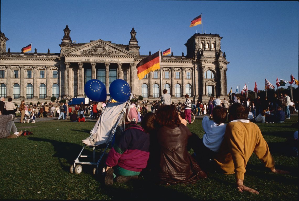Eine Familie mit Buggy an dem zwei Luftballons in den EU-Farben hängen sitzt mit vielen anderen Menschen im Sonnenschein auf dem Rasen vor dem Deutschen Reichstag. Dieser lag bis vor einem Jahr vor der Aufnahme noch in der DDR. Die DDR und die BRD wurden nach dem Fall der Mauer in kürzester Zeit ein gemeinsamer Staat.