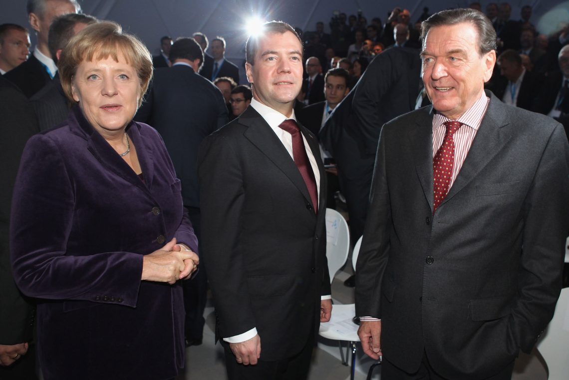 Die frühere Bundeskanzlerin Angela Merkel, der russische Präsident Dmitry Medvedev und der Gazprom-Manager Gerhard Schröder lächeln und blicken Richtung Kamera, aber alle dennoch in eine andere Richtung. Medvedev trägt durch einen Scheinwerfer hinter ihm eine Aurora um seinen Kopf. Das Bild ist Teil eines Beitrags über Russland und die Ukraine und Europa.