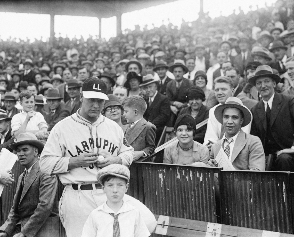 Lou Gehrig von den New York Yankees steht vor einer Tribüne mit vielen Menschen und signiert einen Baseball. Er trägt seine Baseballkleidung und eine Baseballkappe mit LL auf der Stirn. EIn Junge mit Schirmmütze steht vor ihm und blickt in die Kamera.