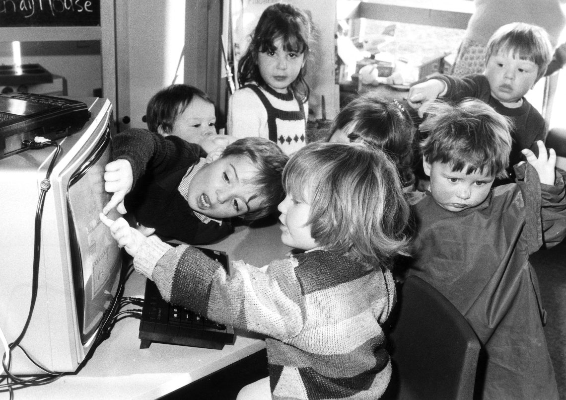Eibe Gruppe von kleinen Kindern ist um einen Computer versammelt und gestikuliert aufgeregt. Ein Mädchen blickt direkt in die Kamera.