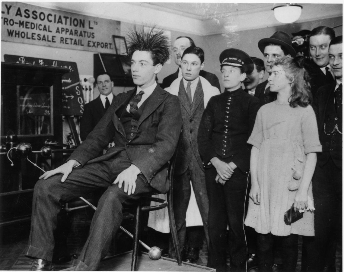 Schwarz-weiß Foto eines Mannes auf einem Stuhl, dem die Haare zu Berge stehen. Er ist von einer Gruppe Menschen umgeben, die zuschauen. Es handelt sich um eine Verkaufsveranstaltung für eine medizinische Prothese im Jahr 1923.