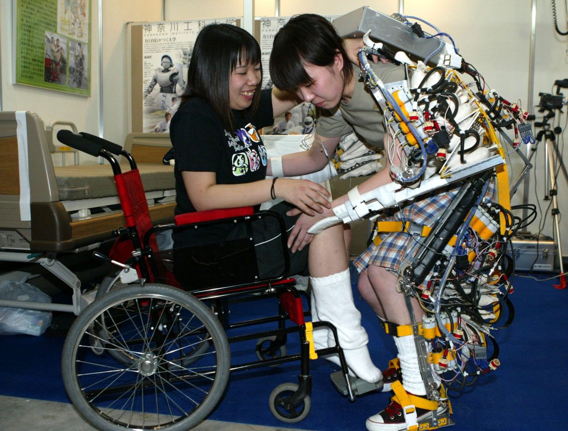 Zwei Frauen, die eine im Rollstuhl, die andere mit einem Exoskelett mit vielen Kabeln, lachen. Die Frau mit Exoskelett versucht sich aufzurichten, was nicht zu gelingen scheint. Das Bild illustriert einen Beitrag über Cyborgs.