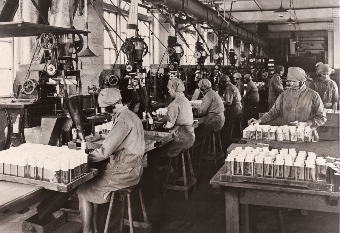 Frauen sitzen auf dem schwarz-weiß Foto an Maschinen in gebückter Haltung, andere stehen an hohen Tischen und zählen Verpackungen. Es handelt sich um einen Arbeitsraum der Rowntree-Fabrik in Yorkshire.