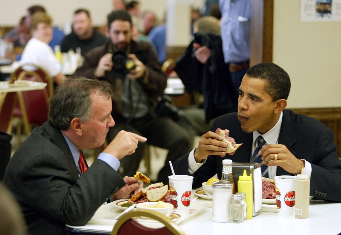 Foto von Barack Obama bei einem Mittagessen mit Richard Daly in Chicago in einem traditionellen Deli. Ein Fotograph im Hintergrund dokumentiert das Treffen. Weitere Gäste des Lokals drehen sich zu der Szene um. Richard Daly deutet mit dem Finger udn Obama scheint überrascht innezuhalten.