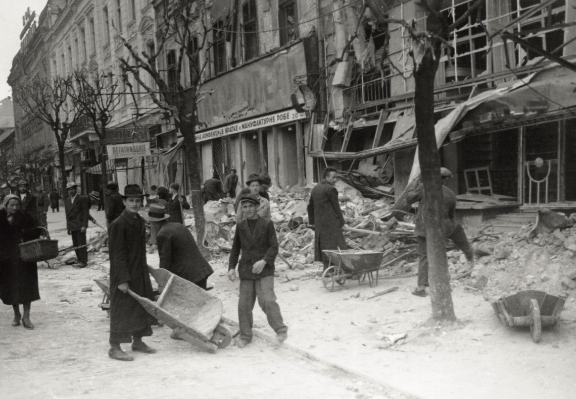 Schwarz-weiß Foto einer Straße mit zerstörten Häusern und kahlen Bäumen. Menschen in Mänteln stehen mit Schubkarren und räumen die Zerstörungen auf. Die Aufnahme stammt aus dem Jahr 1941 in Belgrad. Es sind Kriegsschäden in Folge der Bombardierung durch die deutsche Luftwaffe.