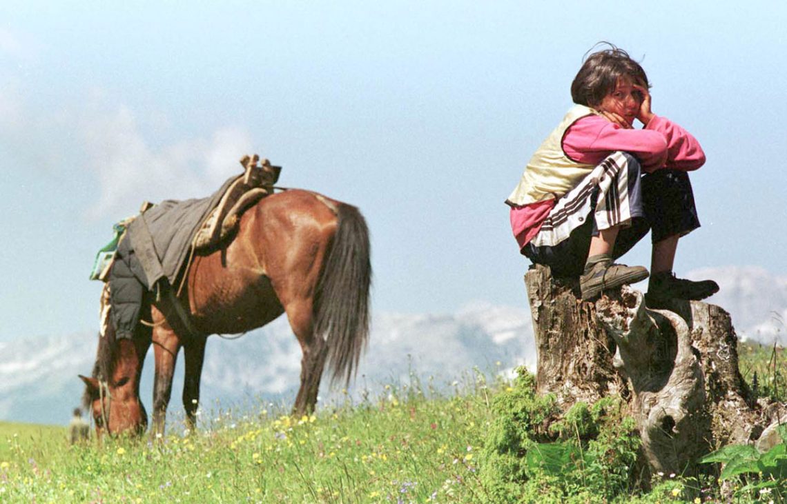 Ein Mädchen hockt auf einem Baumstumpf und blickt in die Kamera. Im Hintergrund steht ein Pferd. Das Bild zeigt die Flucht von Albanern aus dem Kosovo nach Albanien 1998. Die im Kosovo lebenden Albaner wurden von den Serben angegriffen und vertrieben.