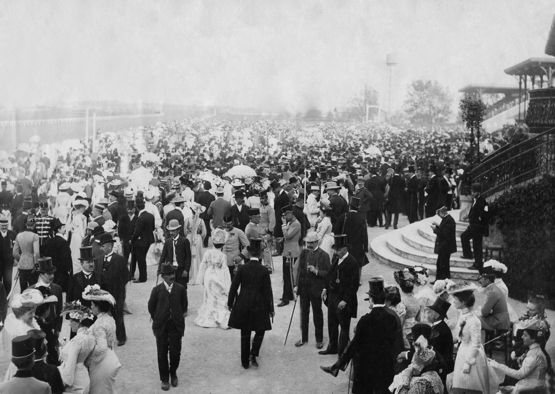 Foto elegant gekleideter Menschen am Rande einer Pferderennbahn mit Jugendstilgebäuden. Die Männer tragen Zylinder und die Frauen große Hüte. Die Aufnahme stammt von 1902 und wurde in Wien gemacht.