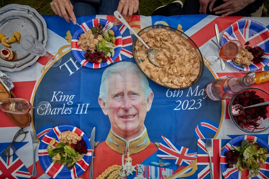 Foto eines gedeckten Tisches aus der Vogelperspektive. Der Tisch ist mit einer Tischdecke mit dem Portraitfoto von Prinz Charles gedeckt. Auf der Tischdecke in den Farben des Union Jack und mit der britischen Flagge steht das Datum 6th May 2023 und King Charles III.