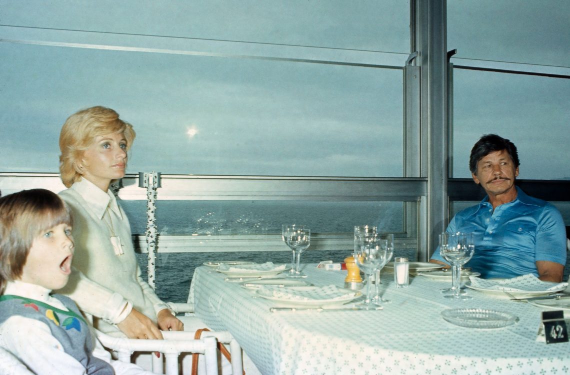 Charles Bronson, Jill Ireland und Zuleika Bronson sitzen an einem mit einem weißen Tischtuch und Servietten im selben Muster gedeckten Tisch mit Tellern und Gläsern, der vor einem großen Fenster mit Aussicht auf ein Meer steht. 