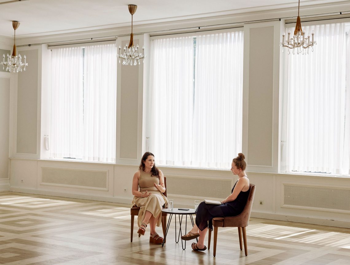 Die gebürtige Ukrainerin Yana Barinova und Stephanie Lehner sitzen einander bei einem Interview gegenüber in einem großen Raum mit Parkettboden und großen Fenstern. Das Bild ist Teil eines Interviews über Kunst in der Ukraine.