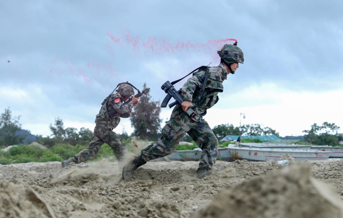 Zwei chinesische Soldaten mit Laser-Tag-Gewehren laufen während einer Übung über einen Sandstreifen. Aus dem Helm des Vordermanns steigt roter Rauch auf, der ihn als ausgeschaltet markiert.
