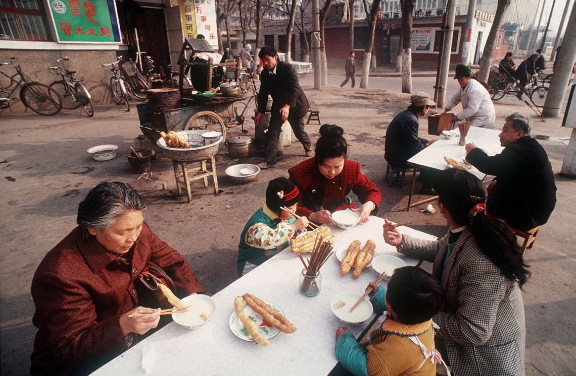 Foto von einem Straßenstand, der frittierte Teigtaschen anbietet, mit einem Tisch mit weißer Resopal-Platte, an dem Kinder, Frauen und Männer sitzen und frühstücken.
