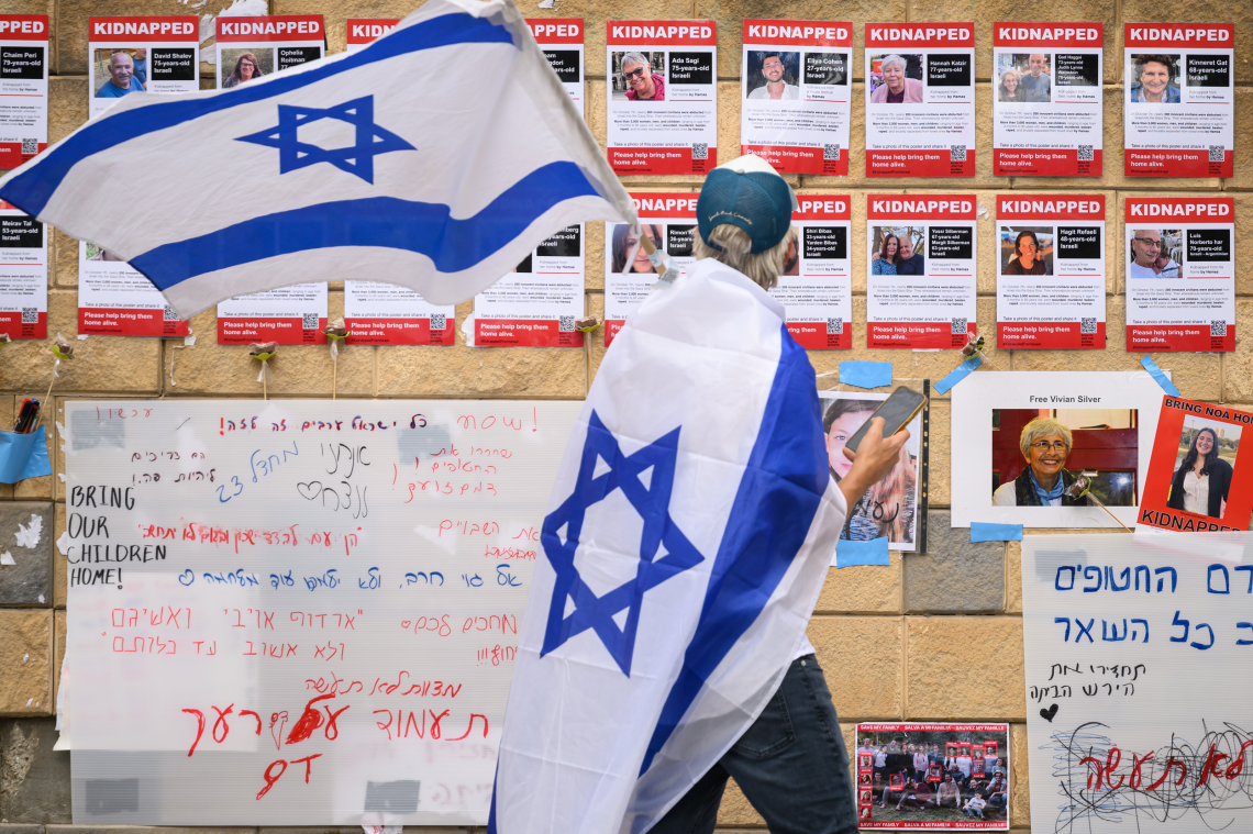 Ein Mann mit Israel-Flagge geht an einer Wand vorüber an der Bilder der Geisel hängen, die von der Hamas entführt wurde.