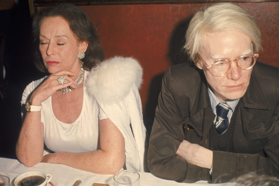Die Schauspielerin Paulette Goddard und der Künstler Andy Warhol sitzen an einem weiß gedeckten Tisch, wobei beide die Arme vor sich verschränkt halten. Goddard trägt große Ohrringe, ein Collier und eine Uhr mit weißem Armband, ein weißes Kleid mit Cape mit Pelzkragen und hat die Augen geschlossen. Warhol blickt von ihr weg zur Seite und trägt ein braunes Sakko mit großen Taschen vorn, einen Pullunder mit V-Ausschnitt udn eine gstreifte Kravatte zu einem hellgrauen oder hellblauen Hemd. Das Bild ist Teil eines Beitrags über Italien und den globalen Erfolg der italienischen Küche. Es illustriert den Podcast macht Hunger mit Peter Peter.