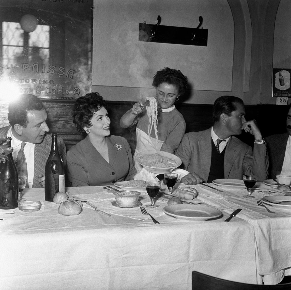 Gina Lollobrigida und ihr Ehemann sitzen an einem Tisch und bekommen grade Spaghetti serviert. Es stehen Wein und Weingläser auf dem Tisch. Die Teller sind einfach und dick. Es liegt auch Brot auf dem Tisch. Die Spaghetti dampfen. Die Serviererin lächelt.