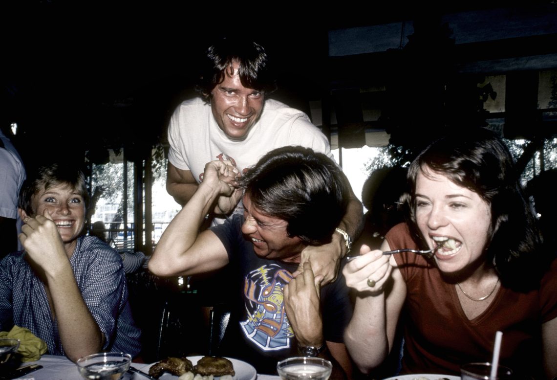 Arnold Schwarzenegger auf einer Aufnahme aus dem August 1977 wie er mit einem Freund kebbelt, wobei der Freund und zwei weitere Freundinnen an einem gedeckten Tisch beim Abendessen sitzen. Arnold Schwarzenegger lacht ebenso wie eine der beiden Frauen in die Kamera, während die andere Frau gerade isst.