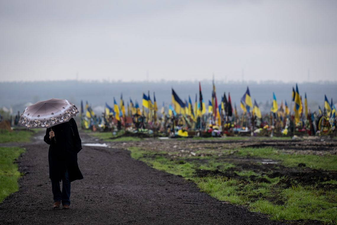 Frau mit Regenschirm. Im Hintergrund markieren ukrainische National- und Militärflaggen, Blumen und Porträts Gräber auf einem weitläufigen Militärfriedhof.