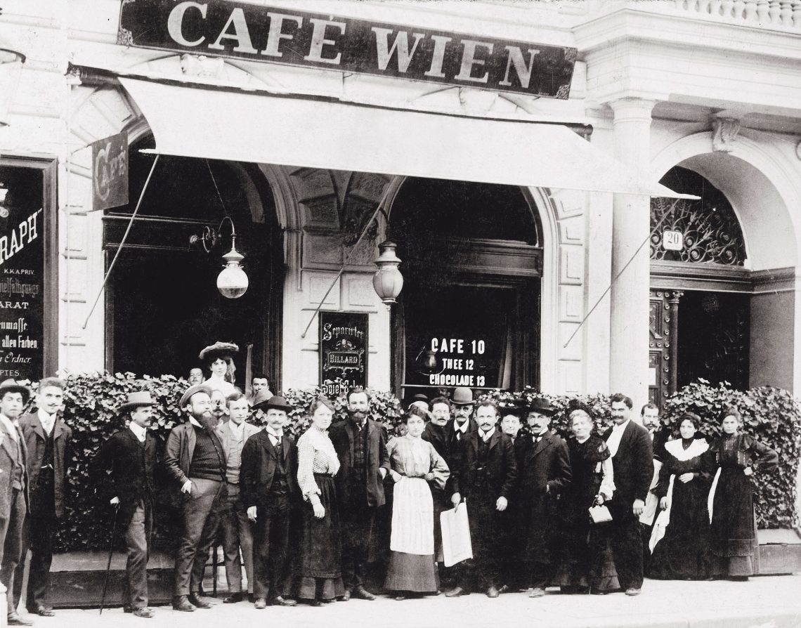Eine Gruppe von Menschen steht vor einem Café das Café Wien heißt. In der Mitte steht eine Frau und hat die Hände in die Hüften gestützt. Das Bild illustriert einen Beitrag über Schnitzel.