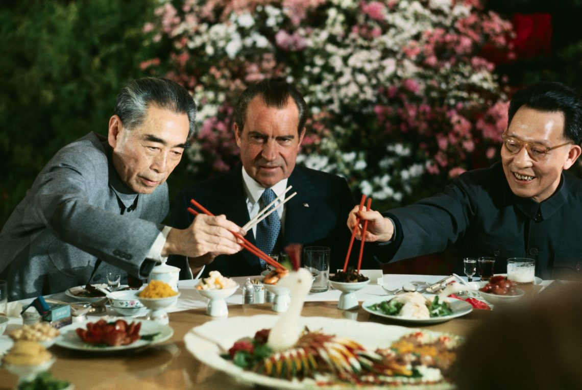 Ein festliches Abendessen. Richard Nixon blickt angespannt und verunsichert auf seine Gastgeber, den chinesischen Premier Zhou Enlai und Chang Chun-chiao von der KP China. Im HIntergrund ein großes Blumenbouquet. Das Bild illustriert einen Beitrag über China und die Gefahr eines Krieges und die Bedeutung für Europa.
