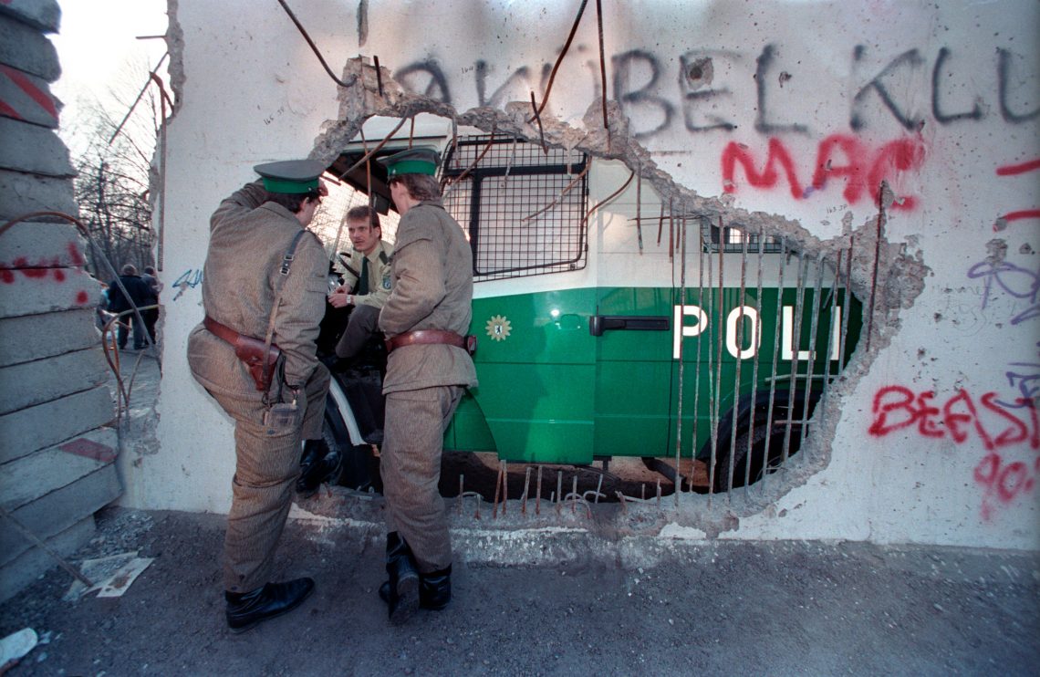 Zwei Polizisten der DDR stehen an einem Rest der Mauer und sprechen mit einem Polizisten der BRD der auf der anderen Seite des Lochs in der Mauer am Lenkrad eines Polizeibuses sitzt. Das Bild illustriert einen Beitrag über die slawische Küche, da eine These Beitrags ist, das die Ost-West-Grenze auf kulinarischer Ebene nach dem Mauerfall noch undurchlässiger war.
