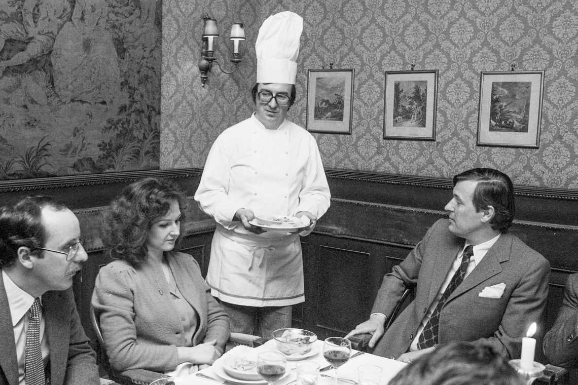 Ewald Plachutta mit Kochhaube steht, Teller in der Hand, bei einem Tisch und blickt auf seine Gäste. Alle schauen ernst. Das Bild illustriert einen Podcast.