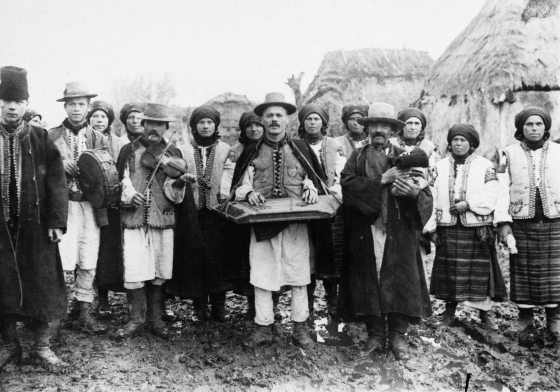 Schwarz-weiß Foto einer Hochzeitsgesellschaft in traditioneller Kleidung. Die Männer tragen Westen mit Stickereien und Hüte; die Frauen ebenfalls die Westen und ein Tuch um den Kopf gewickelt sowie gesteifte Röcke. Alle blicken ernst in die Kamera. Die Aufnahme stammt aus dem ahr 1920.