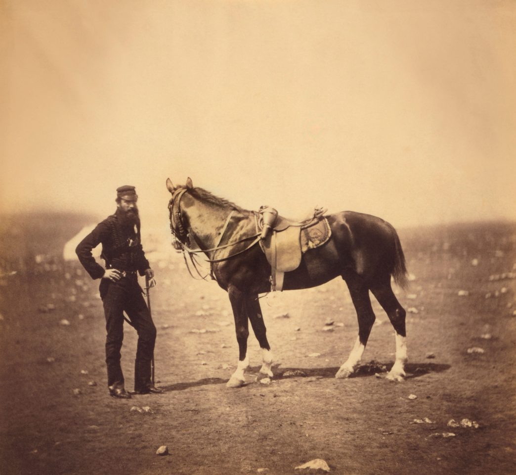 Sepia-Foto eines Mannes in Uniform, der vor einem Pferd steht, das zur Seite blickt. Das Bild wurde während des Krim-Krieges (1853 bis 1856) gemacht.