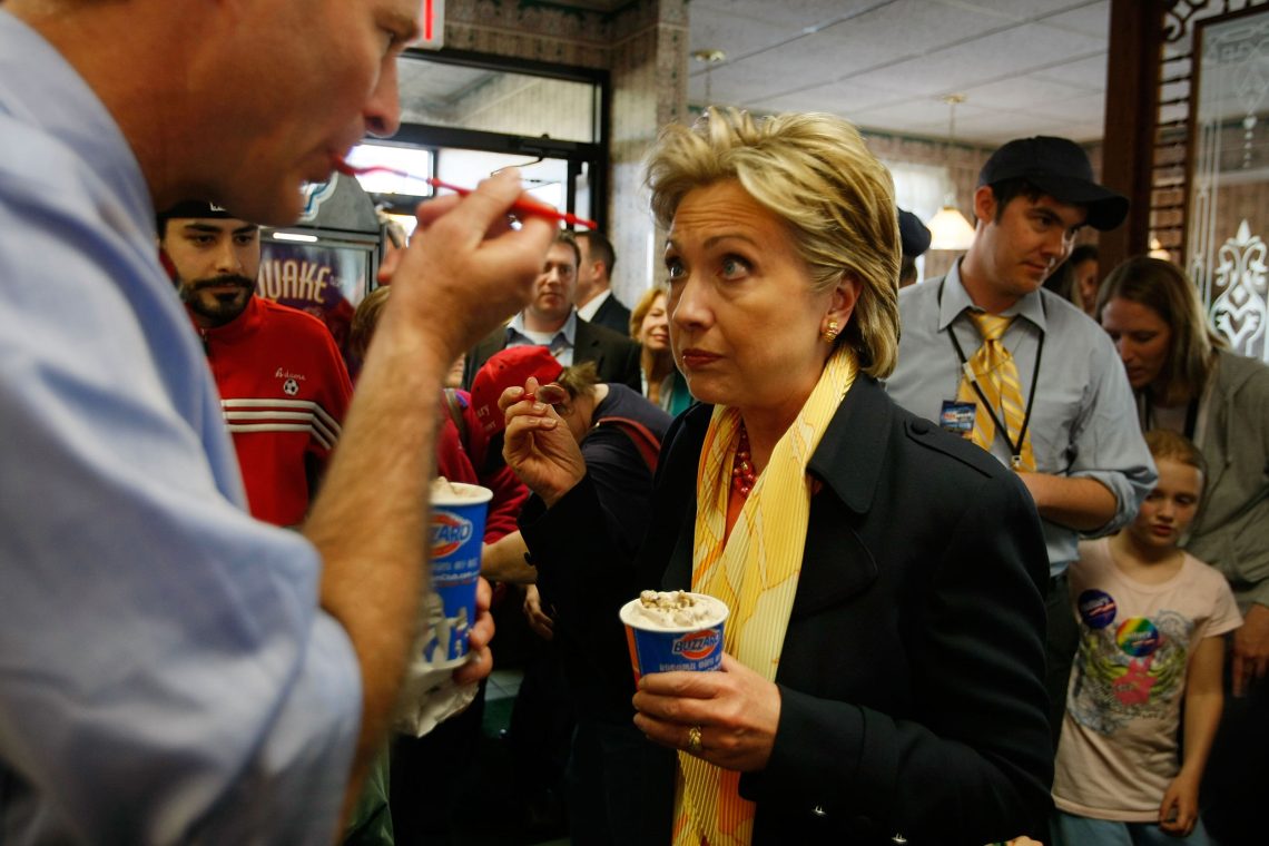 Hilary Clinton isst aus einem Pappbecher eine Süßigkeit und blickt zweifelnd auf einen Mann, der ebenfalls aus einem Pappbecher die Süßigkeit ist.