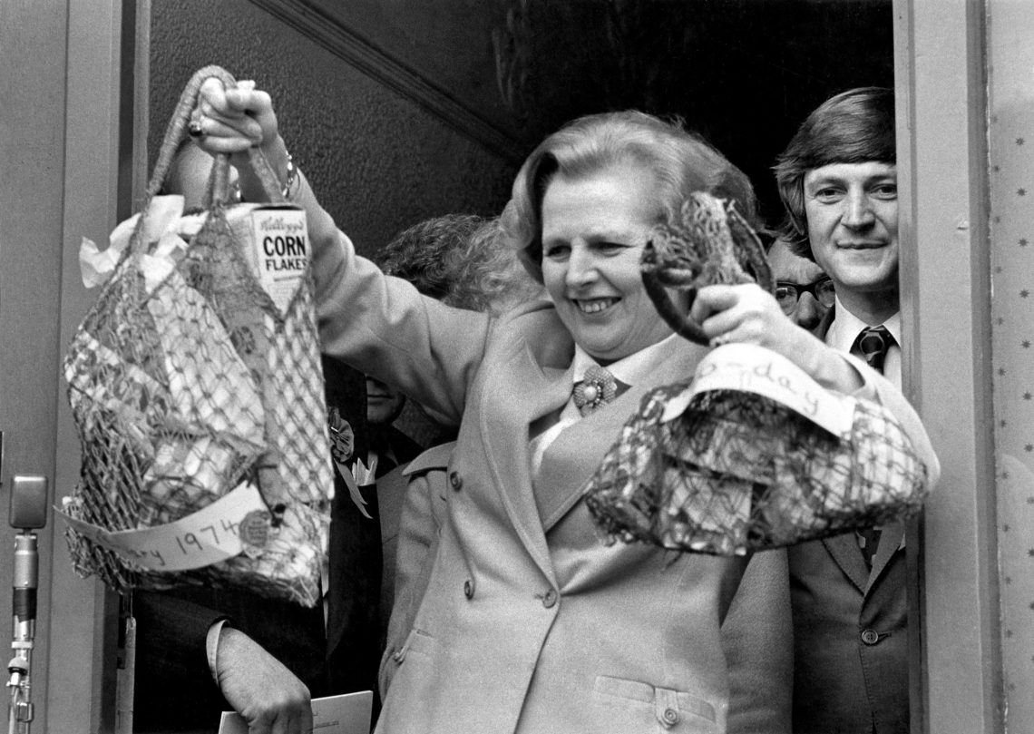 Margaret Thatcher hält bei einer Wahlkampfveranstaltung 1979 triumphierend zwei Einkaufsnetze in die Höhe. Der eine ist wenig befüllt, der andere reichlich. Hinter ihr steht eine Reihe von Menschen. Thatcher tritt gerade aus der Tür. Das Bild illustriert einen Beitrag über Inflation und Zucker.