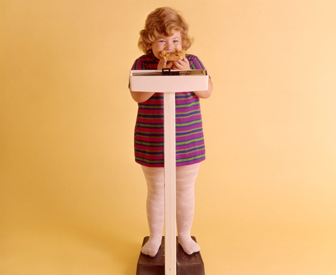 Ein kleines Mädchen steht auf einer Waage, während es ein Sandwich mit Erdnussbutter isst. Das Bild illustriert einen Artikel zum Thema Prävention bei übergewichtigen Kindern.