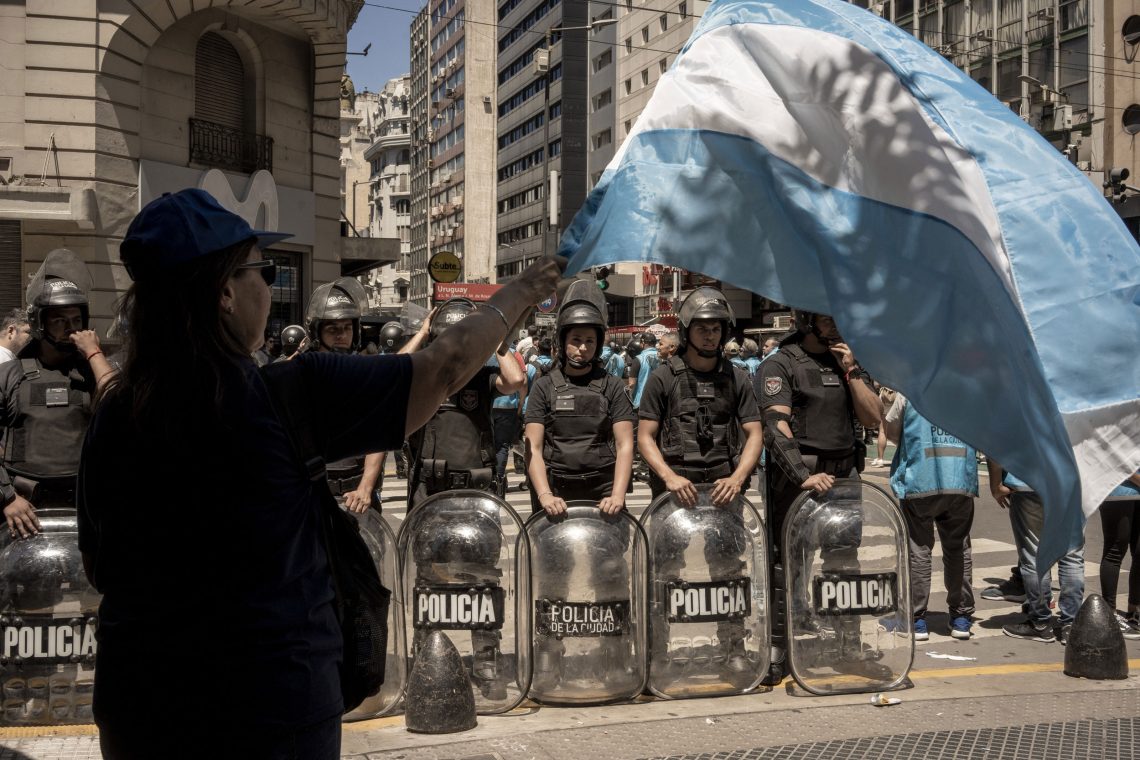 Eine Frau schwenkt eine argentinische Fahne während Polizisten mit Schutzschilden im Hintergrund stehen.