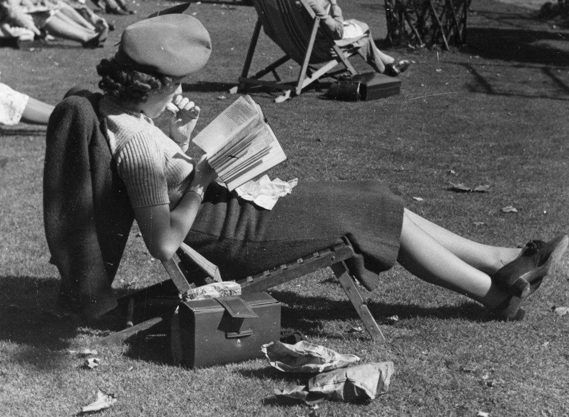 Eine Frau sitzt in einem Liegestuhl in einem Park und liest konzentriert in einem Buch.
